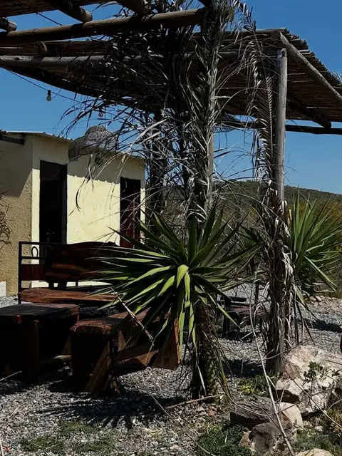 Mesas de madera entre agaves bajo la pérgola, con las sierras de fondo